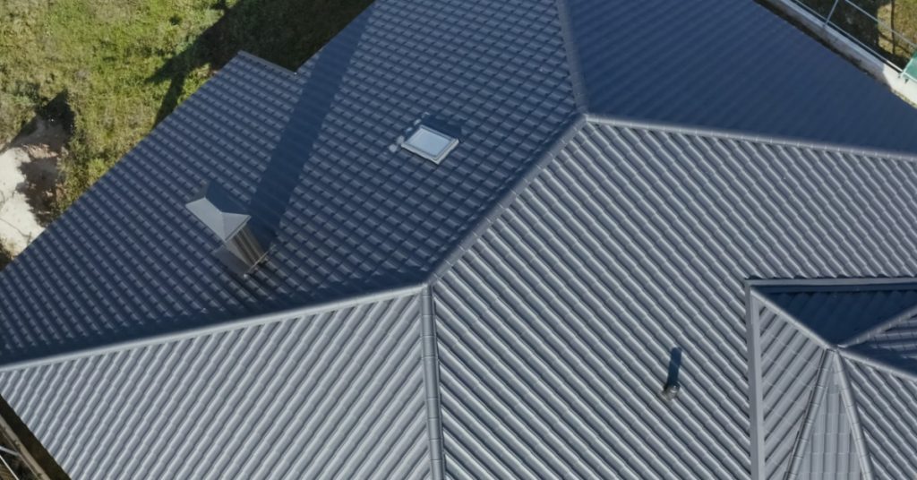 An aerial view of the roof of a home with metal grey shingles and green grass in the front on a sunny day.