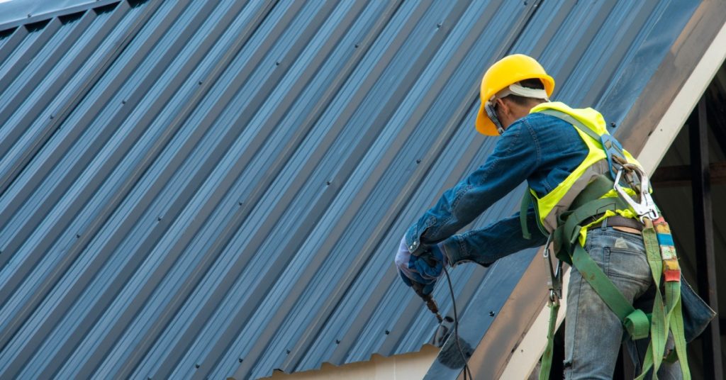 A worker wearing a safety harness and a yellow helmet, using a pneumatic nail gun to attach metal shingles to a roof.