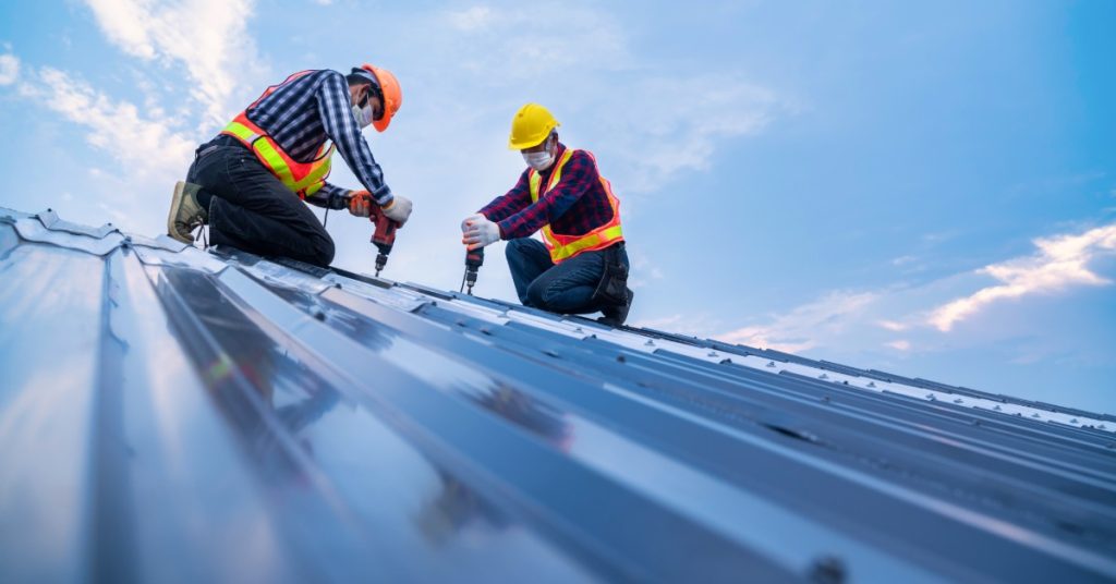 Two workers wearing yellow helmets, kneeling on top of a roof using nail guns to attach metal shingles on a cloudy day.