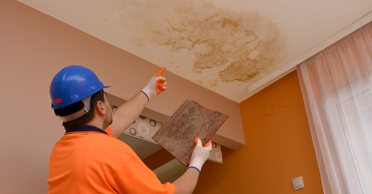 A man wearing an orange shirt and a blue helmet, pointing at a large brown stain in the ceiling of a room.