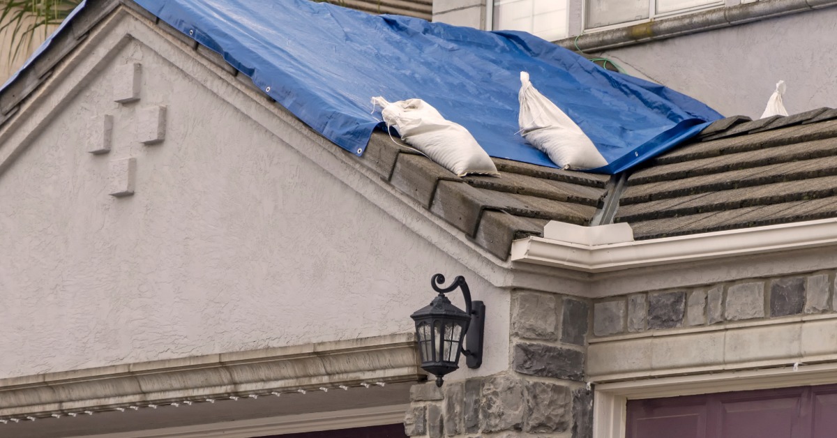 A close-up of the lower roof of a two-story house with brown shingles covered with a large blue tarp held by sandbags.