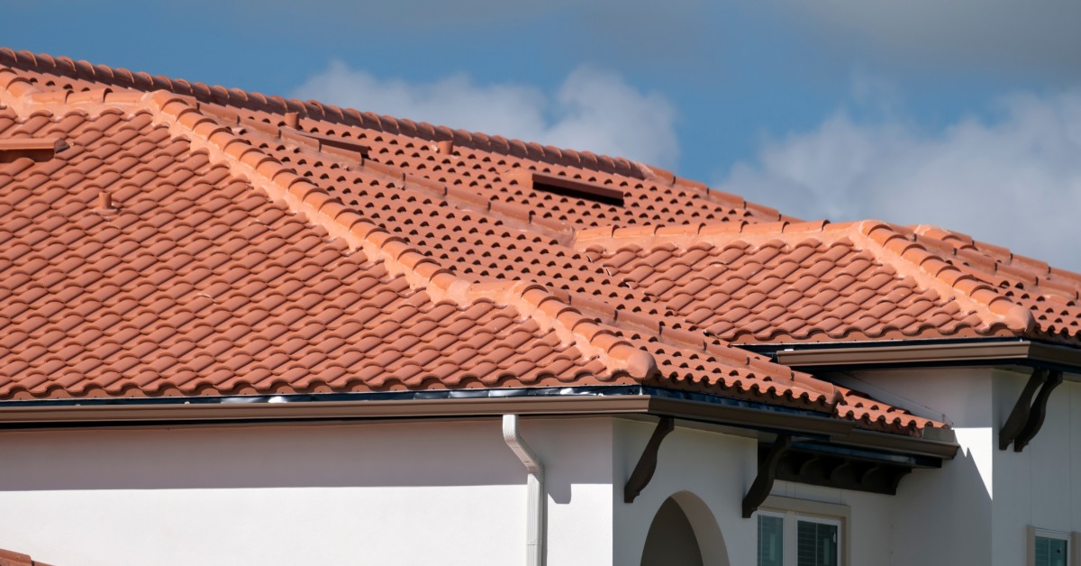 A close-up of the roof of a two-story white home covered in orange clay shingles with some clouds in the background.
