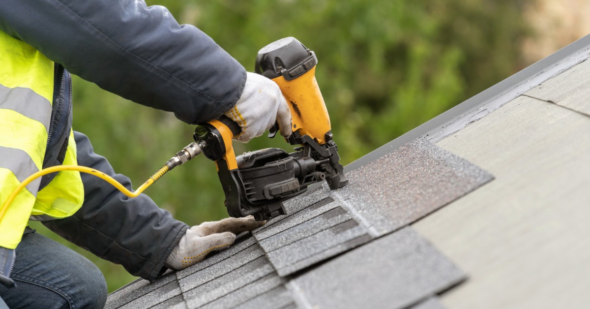 A close-up of the hands of a worker wearing safety gloves and using a pneumatic nail gun to attach roof shingles.