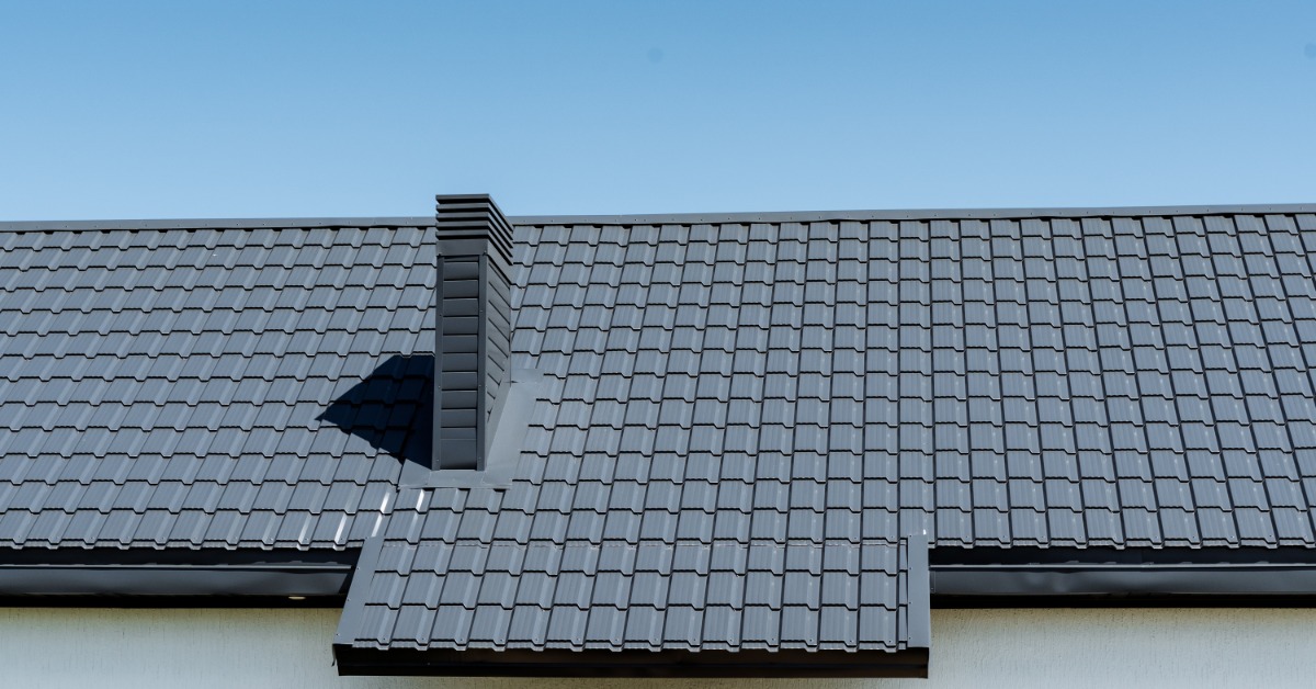 A residential roof with a small chimney covered in grey composite shingles with a blue sky in the background.