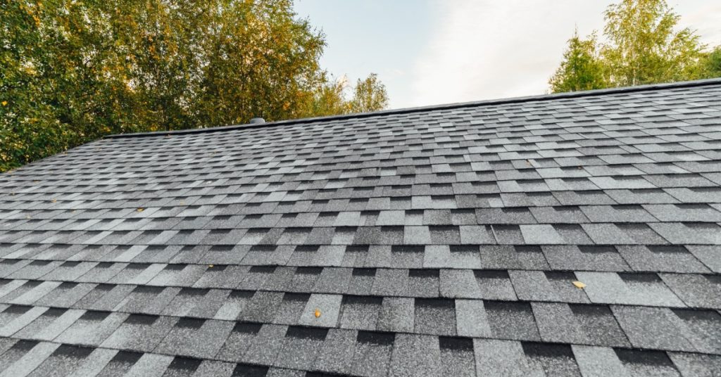 A close-up of a residential roof with grey asphalt shingles of different tones with trees behind on a sunny day.