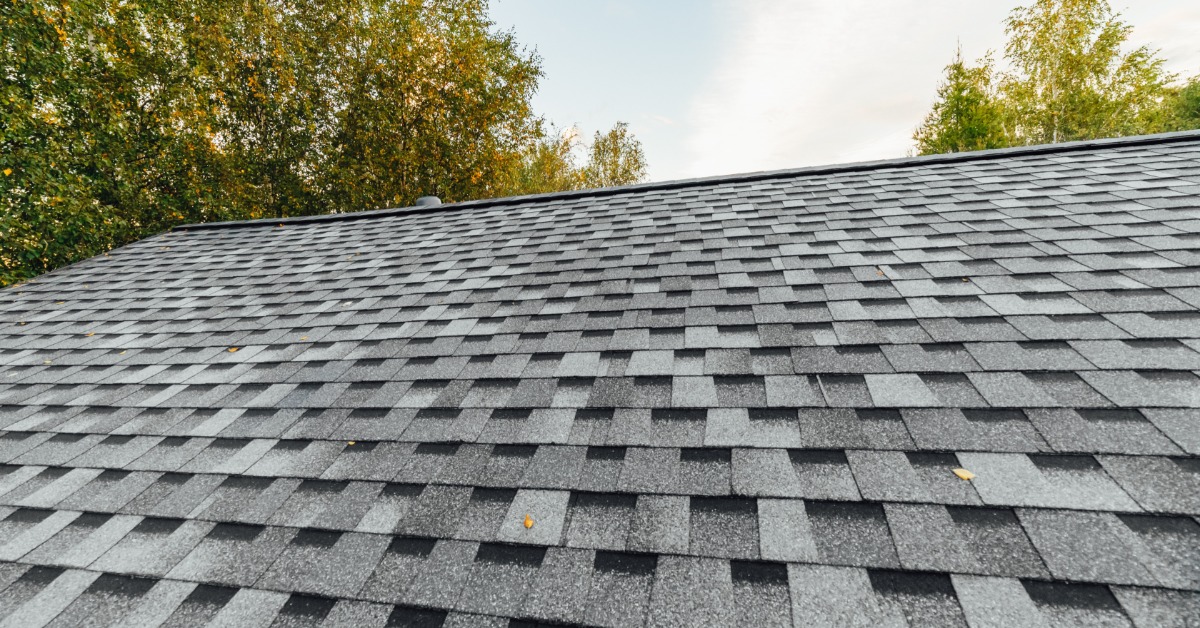 A close-up of a residential roof with grey asphalt shingles of different tones with trees behind on a sunny day.