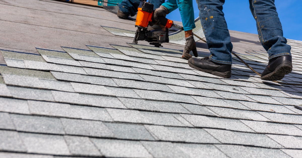 A close-up of a worker wearing gloves and using a pneumatic nail gun to attach asphalt shingles to the roof of a home.