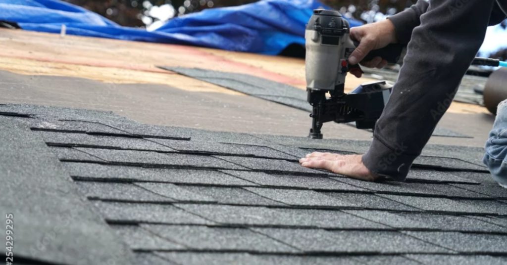 A close-up of two hands using a pneumatic nail gun to attach a grey, asphalt shingle to the roof of a home.