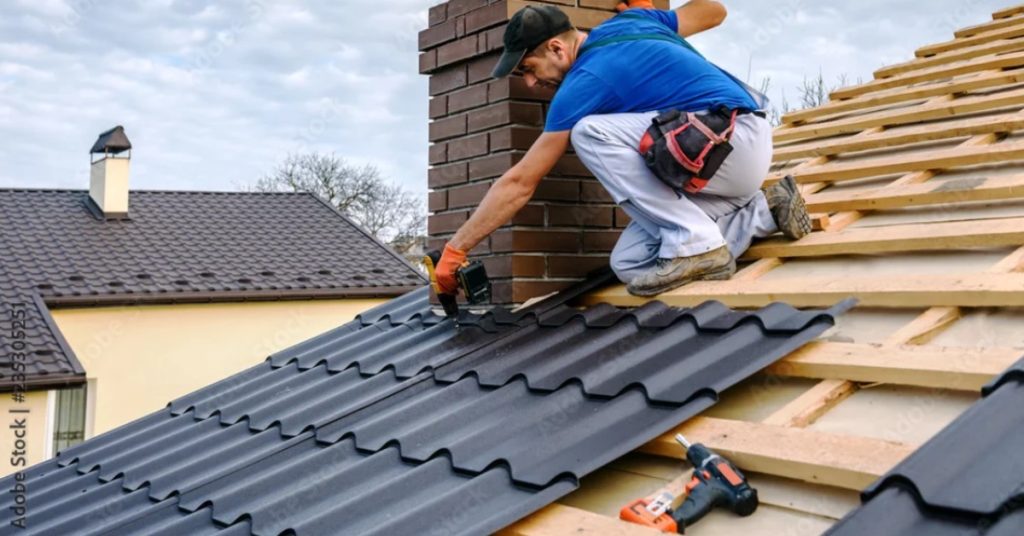 A worker holding on to the brick chimney of a home to attach metal shingles to the roof with an electric screwdriver.