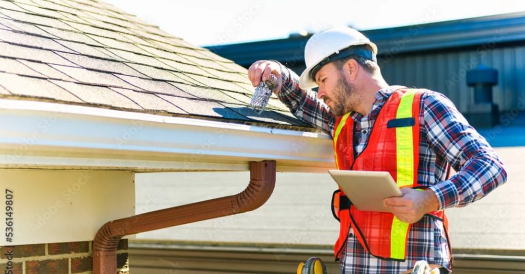 A worker wearing a safety vest and a white helmet looking up close to a shingle on the edge of a residential roof.