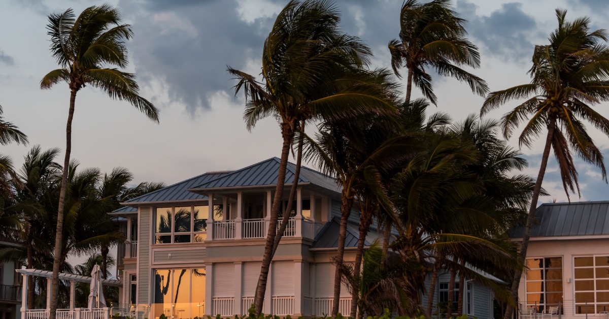 A large two-story home with a metal roof surrounded by multiple palm trees moving with the wind under a cloudy sky.