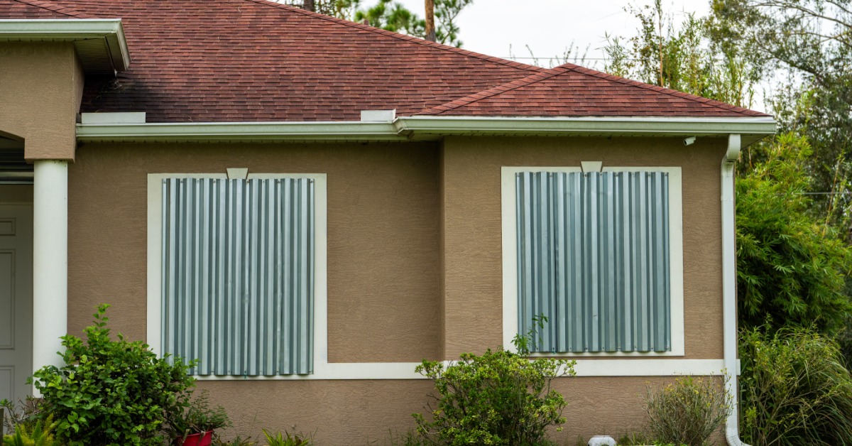 A one-story home with a red roof and sealed-off windows surrounded by greenery and dark clouds in the sky.