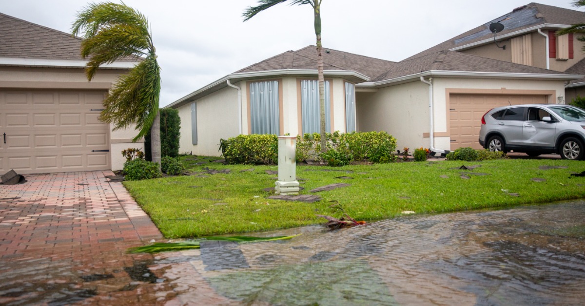 A one-story home with a shingles roof and a palm tree in the front, surrounded by water with multiple clouds above.