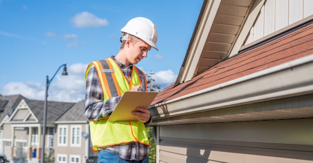 A worker wearing a safety vest and a white helmet standing on top of a ladder while closely looking at roof shingles.