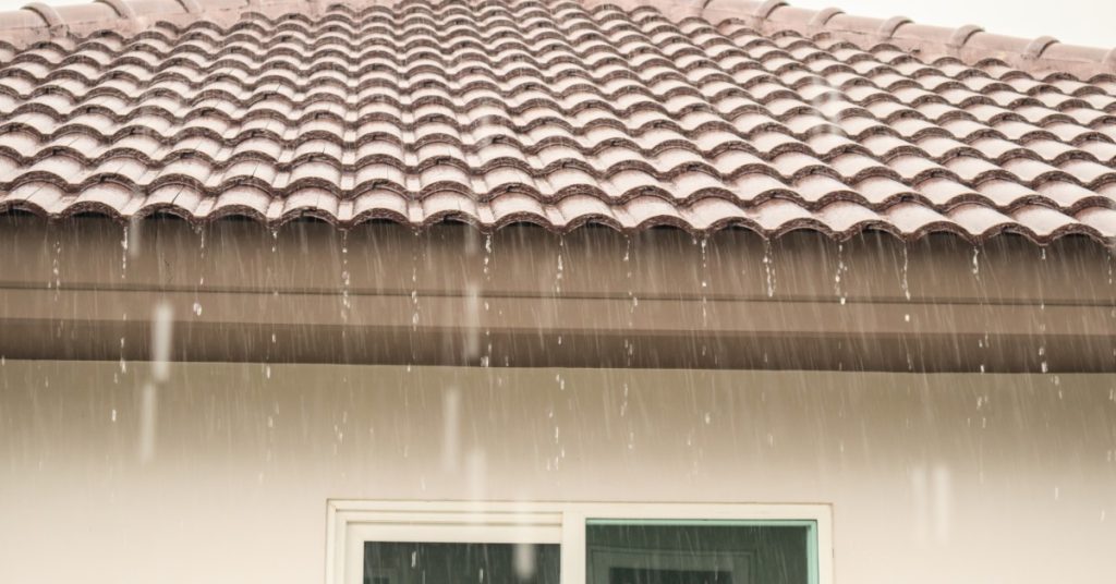 The roof of a home covered in clay shingles with water pouring down from the edges, with black clouds in the background.
