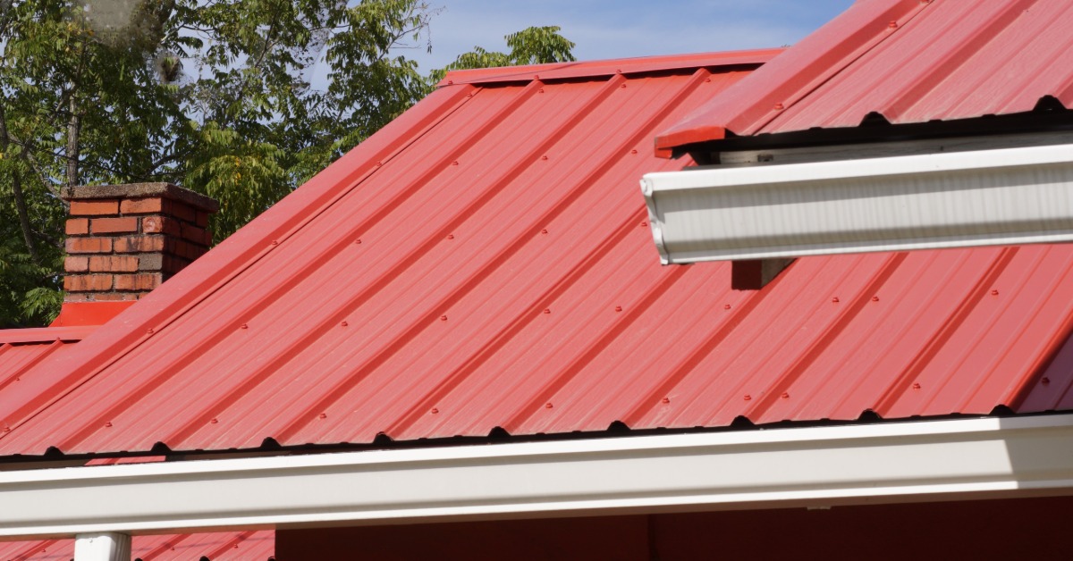 A red metal roof next to a white gutter over a house with red walls and a brick chimney with trees in the background.
