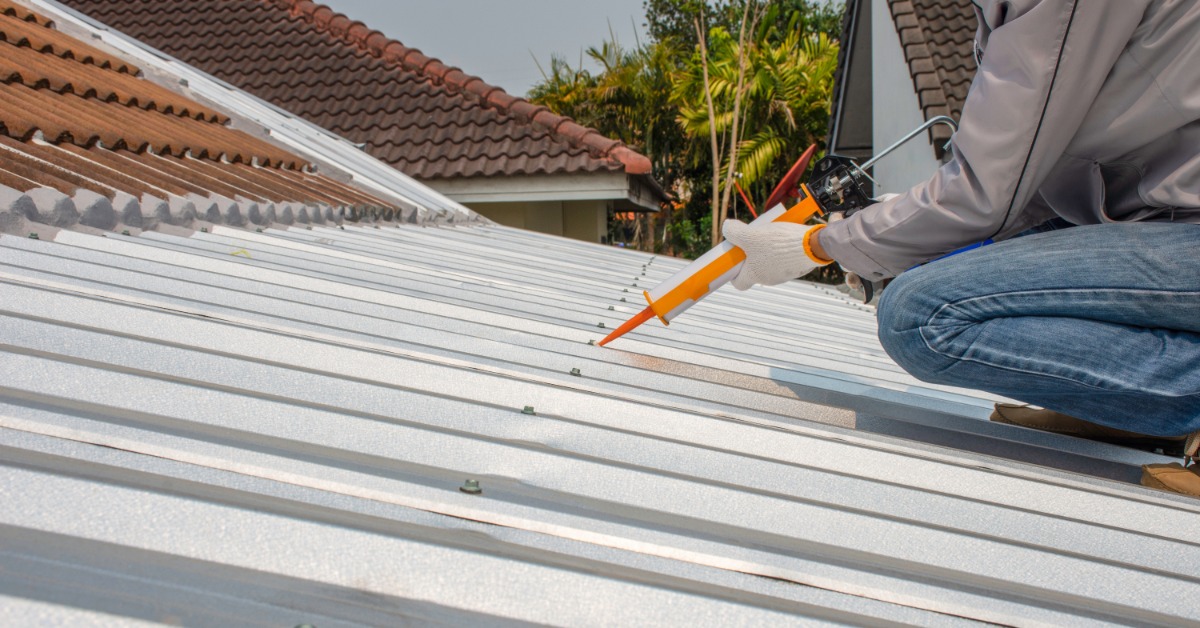 A worker wearing white gloves using a glue gun on the metal roof of a home next to a section with clay tiles.
