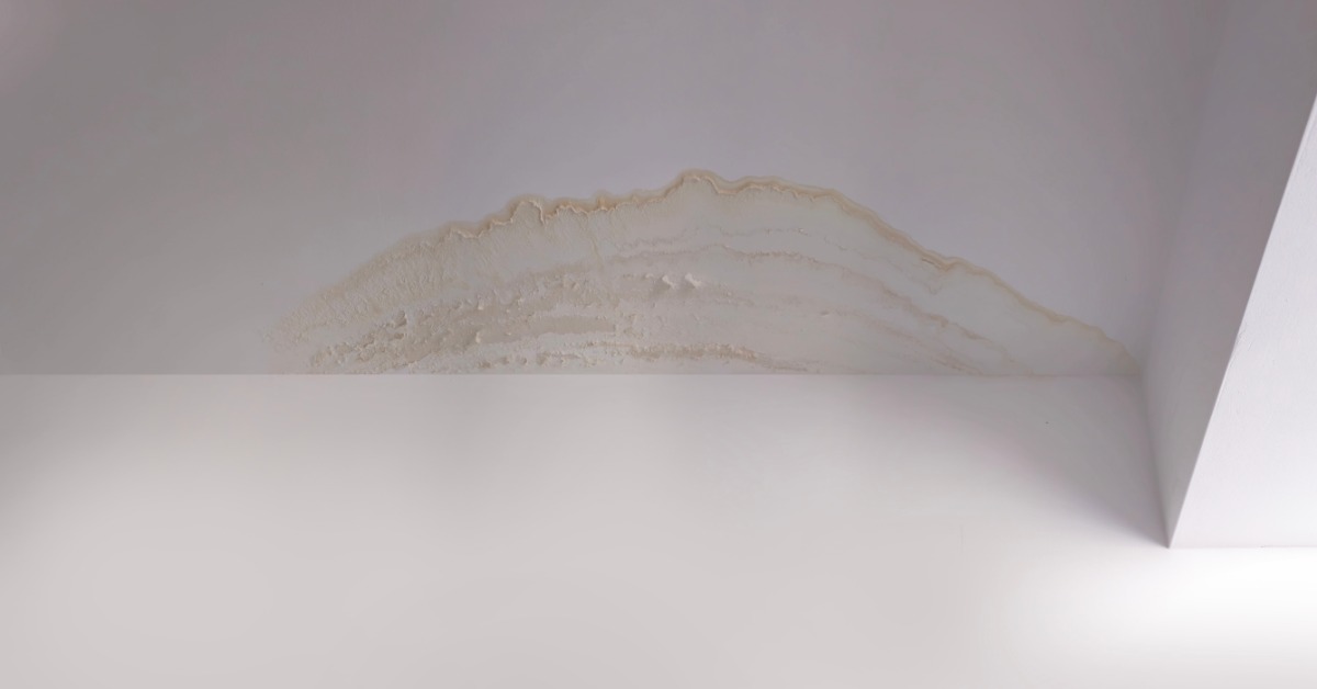 A light yellow water stain on the corner of a white ceiling next to a white wall in a room with grey metal doors.